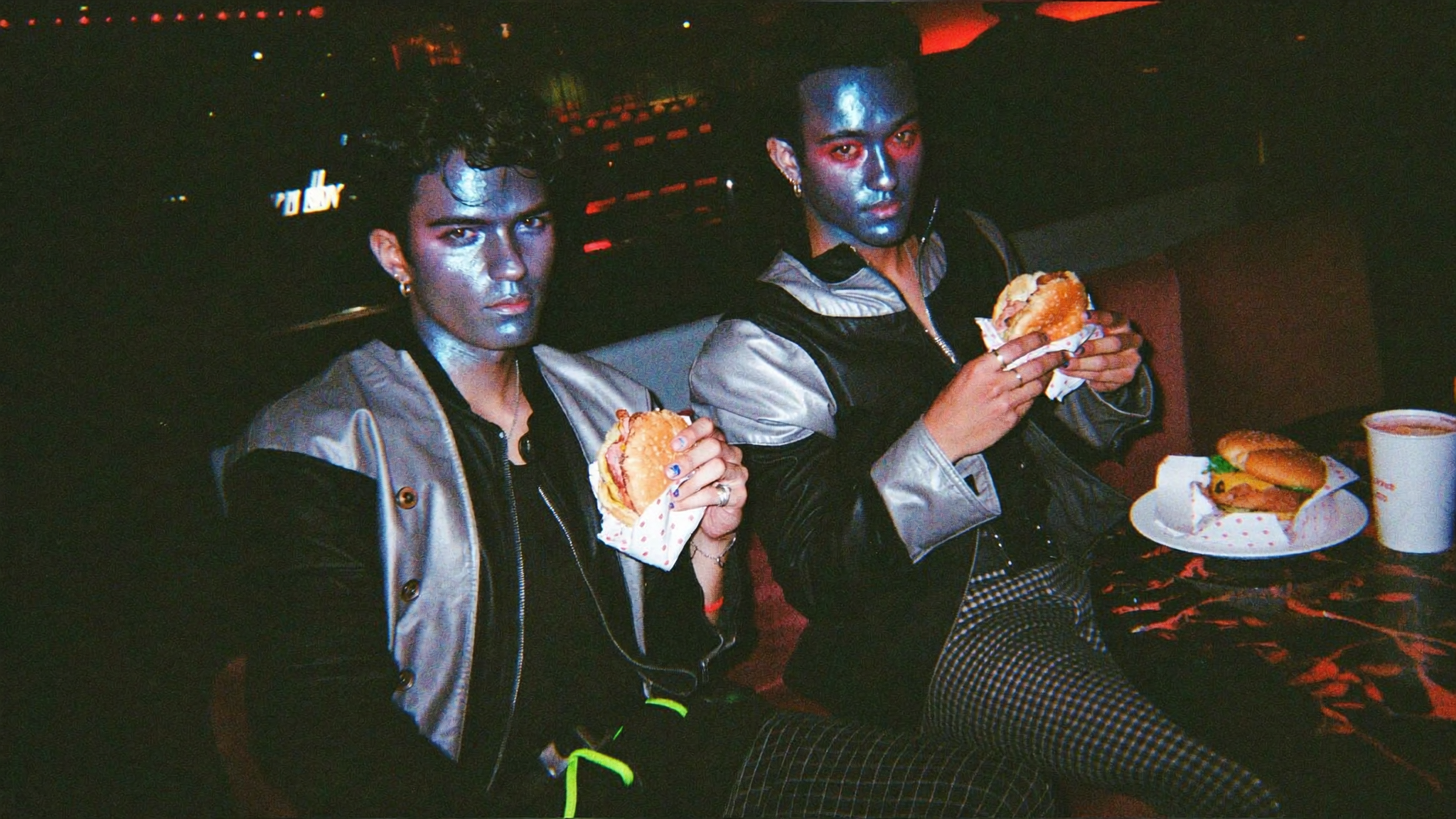 Two silver-faced figures eating hamburgers in a club — the work made in public with dirty hands